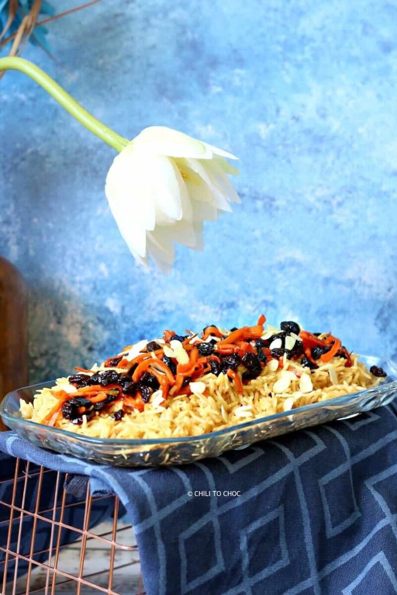 Rice with sliced carrots and raisins on a translucent platter with a tea towel underneath it and white flower in the background.