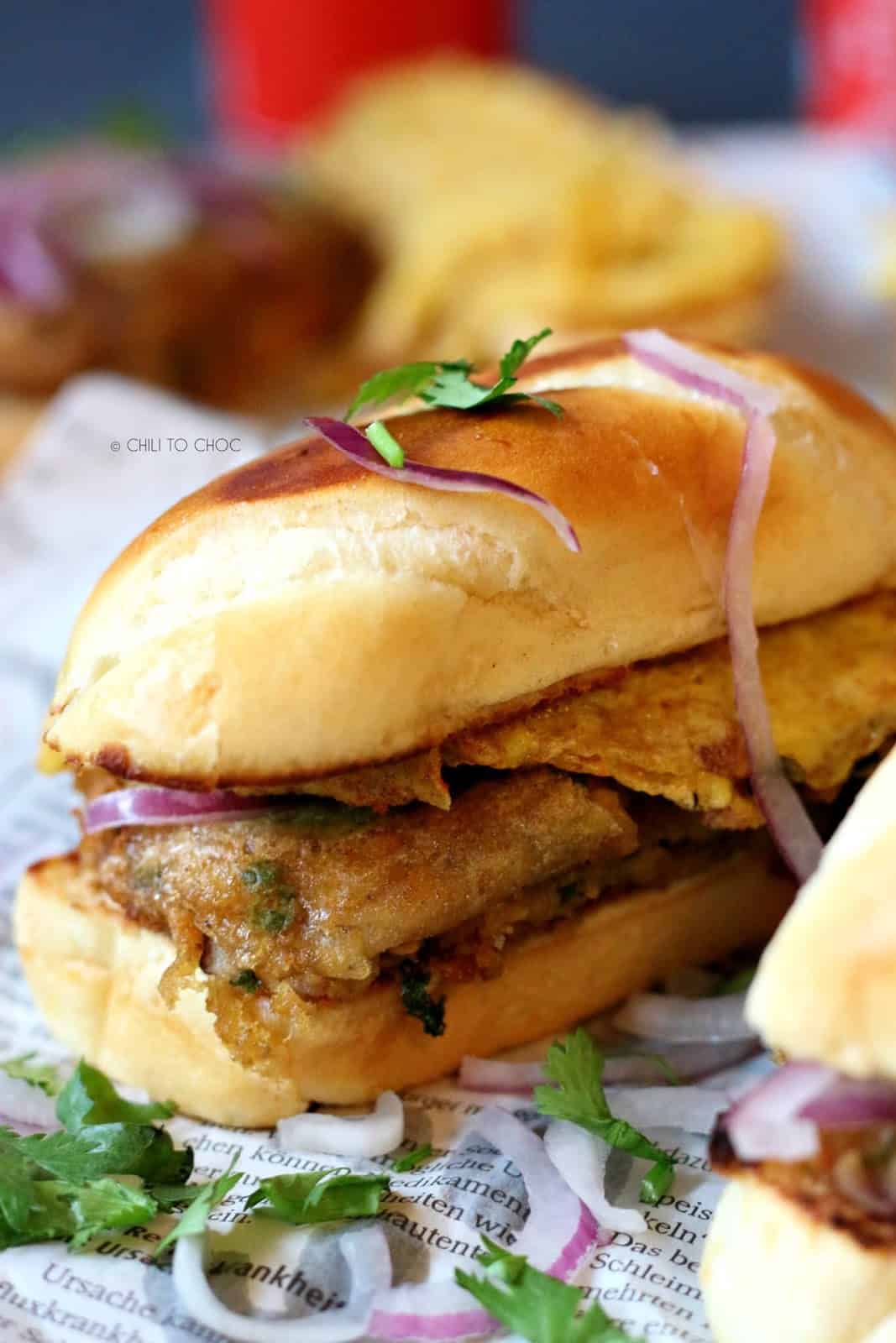 Pakistani Bun Kabab A vegetarian sandwich on a newspaper garnished with red onions and coriander