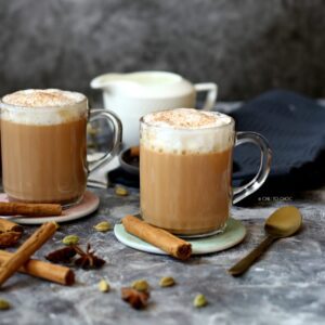 Two clear mugs of chai latte with the whole spices scattered around and a milk pot at the back