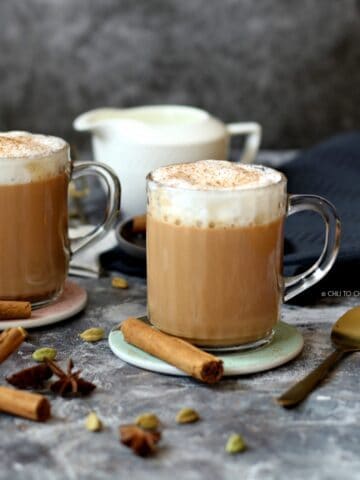 Two clear mugs of chai latte with the whole spices scattered around and a milk pot at the back