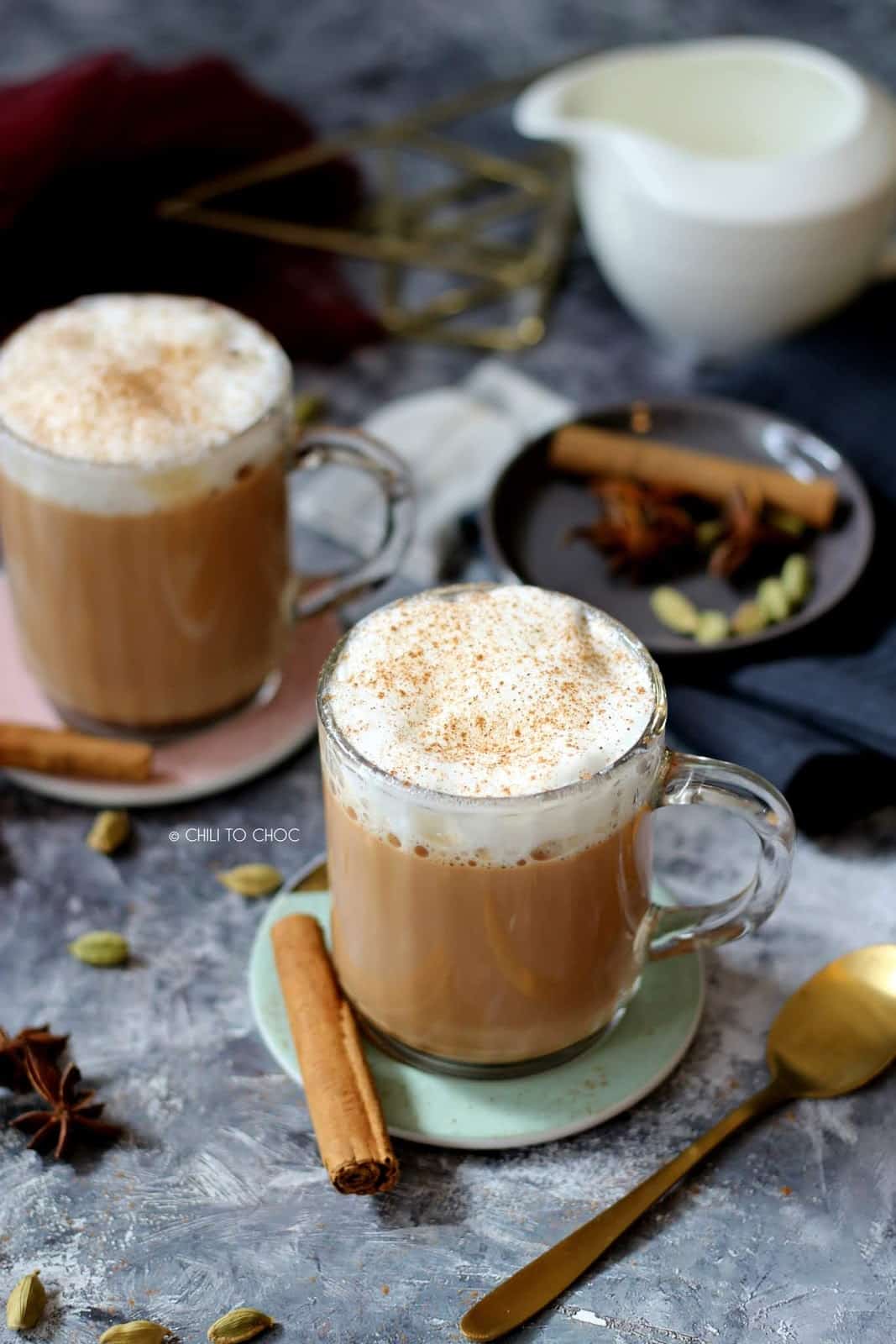 Two mugs of chai tea latte dusted with cinnamon powder and whole spices in the background.