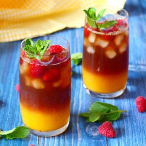 Two tall glasses of mango iced tea garnished with raspberries and mint.