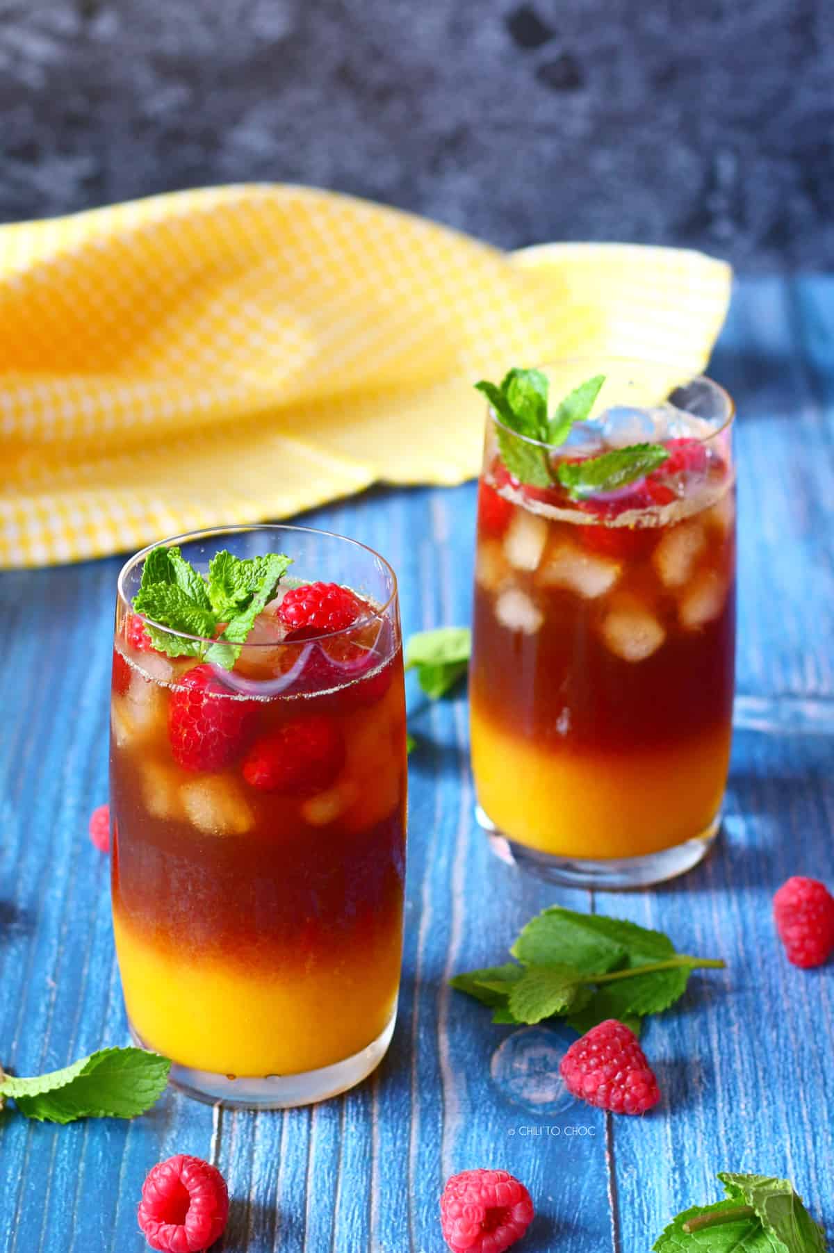 Mango Iced Tea Two glasses of mango iced tea with ice cubes, raspberries and mint on a blue surface.