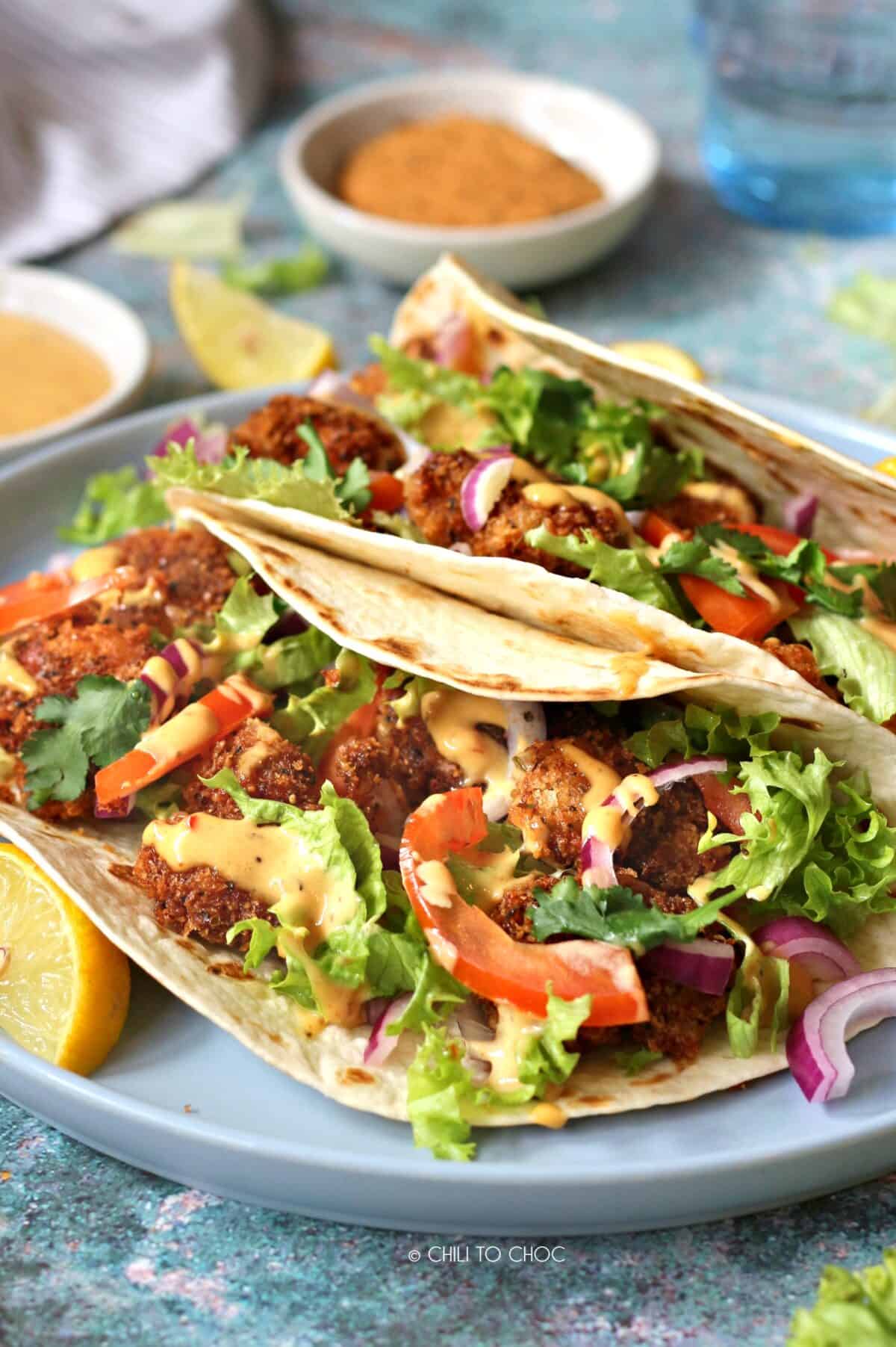 Two fried shrimp tacos on a plate with sliced vegetables and lemon wedges
