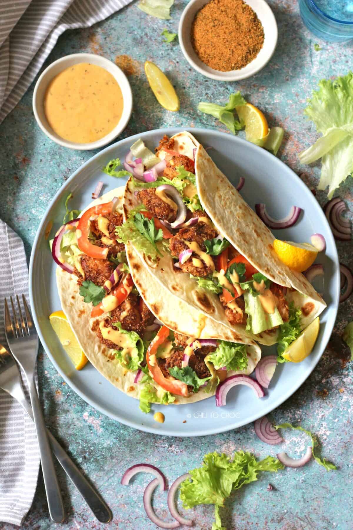 Top shot of two shrimp tacos on a plate with forks on the side and dressing and cajun seasoning on top