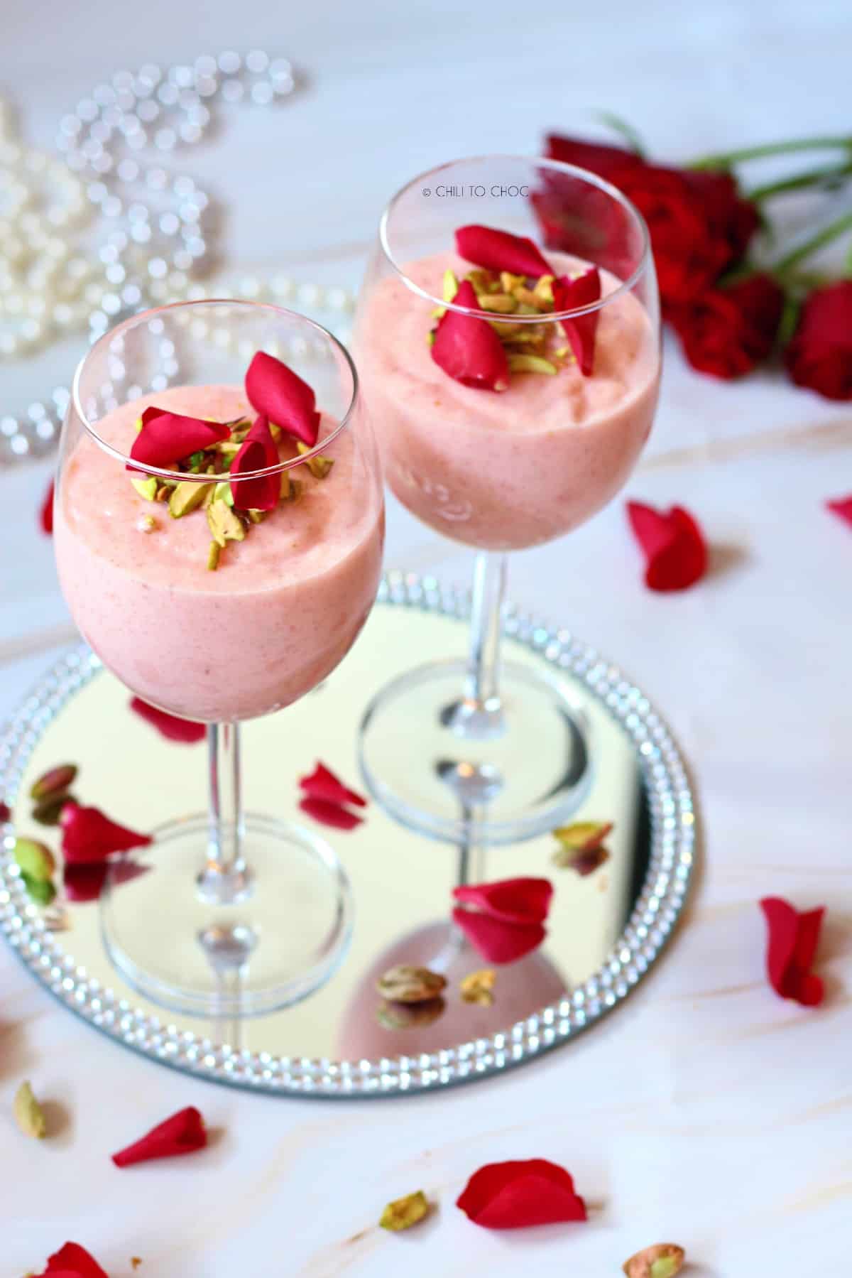 Two stem glasses of pink firni with rose petals on a mirror plate.