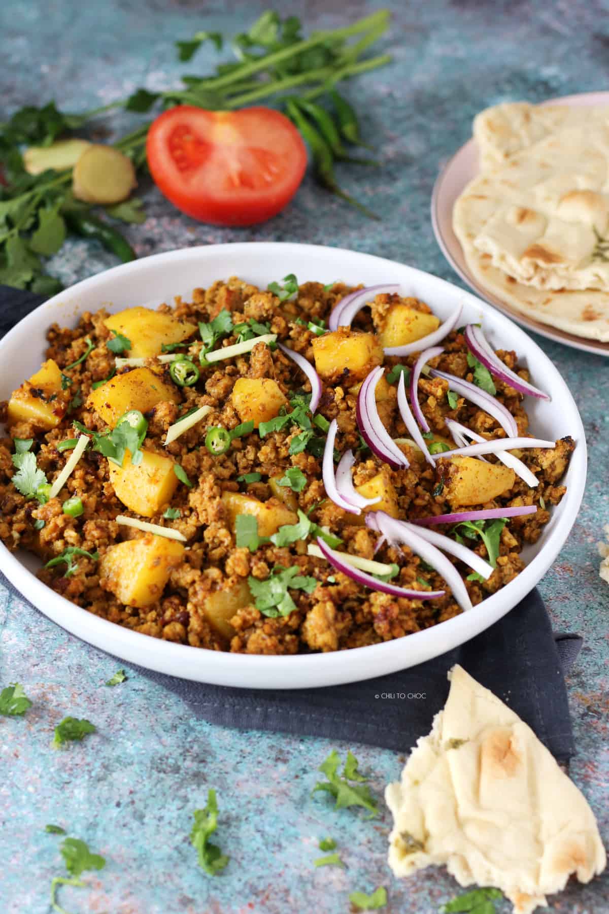 Aloo Keema with garnishes in a white dish and pieces of roti around it.