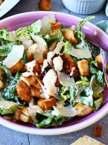 Caesar Salad with grilled chicken, croutons, Parmesan shavings, Caesar dressing in a shallow plate with a fork on the side.