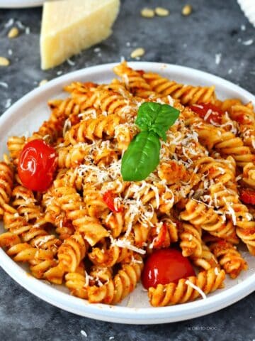 Pesto rosso pasta on a plate garnished with basil leaf and grated Parmesan cheese and a fork on the side.