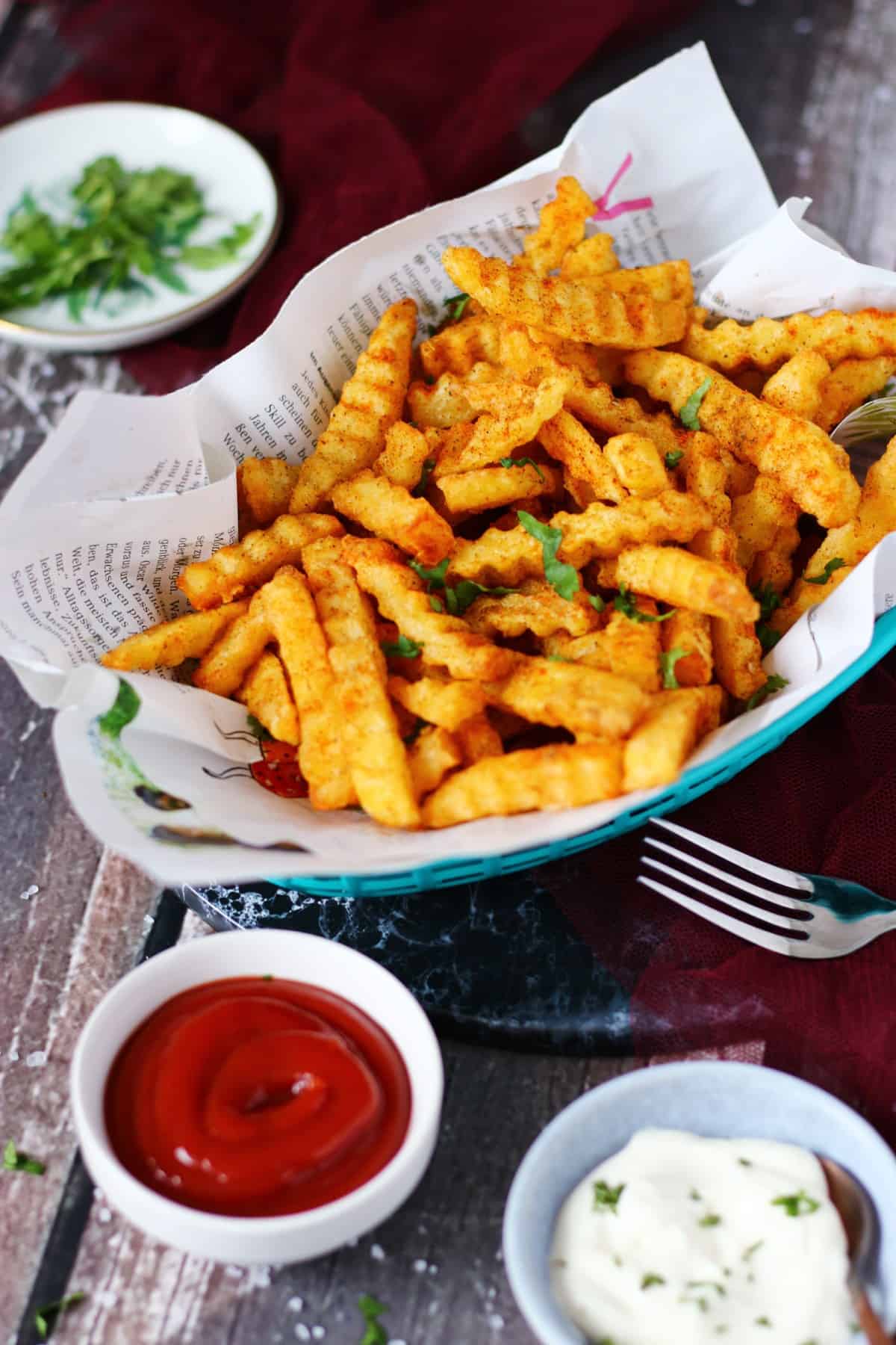 Masala fries in a basket lined with magazine paper and ketchup and mayo bowls on the front.