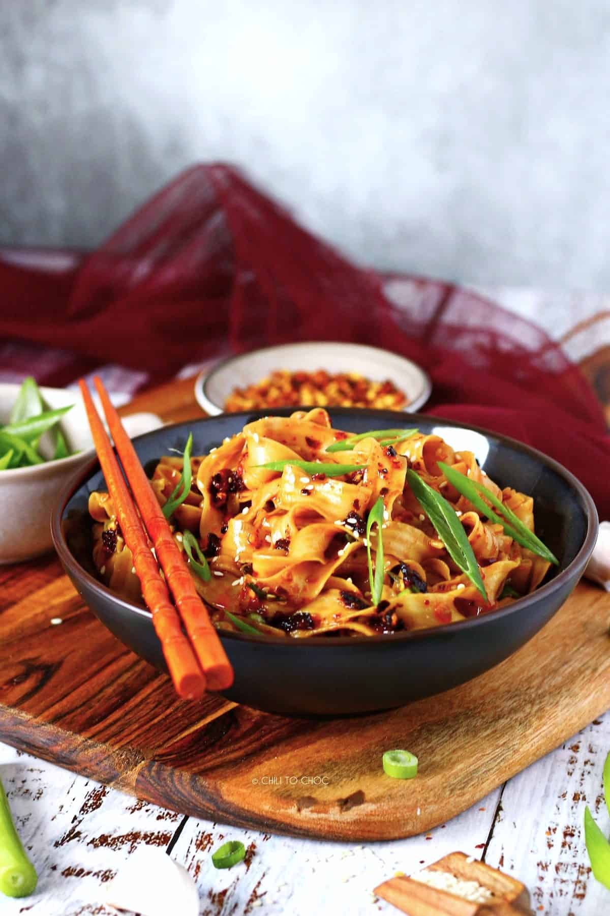 A bowl of spicy noodles on a wooden board with chopsticks on the side.