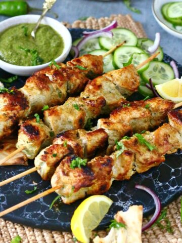 Chicken Malai Boti skewers with green chutney and garnishes on a black marble dish.