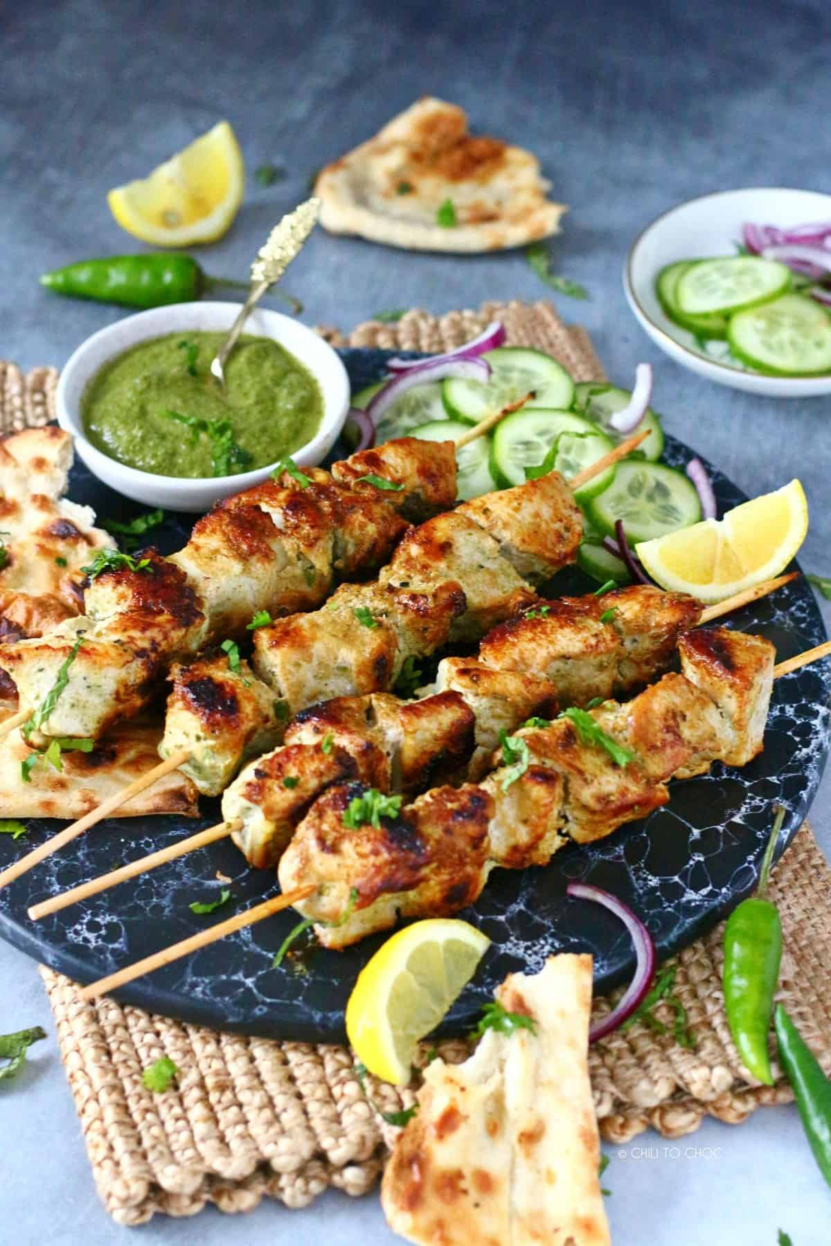 Chicken Malai Boti skewers on a marble dish surrounded by sliced red onion, green chilies, lemon wedges, sliced cucumber and a green chutney.