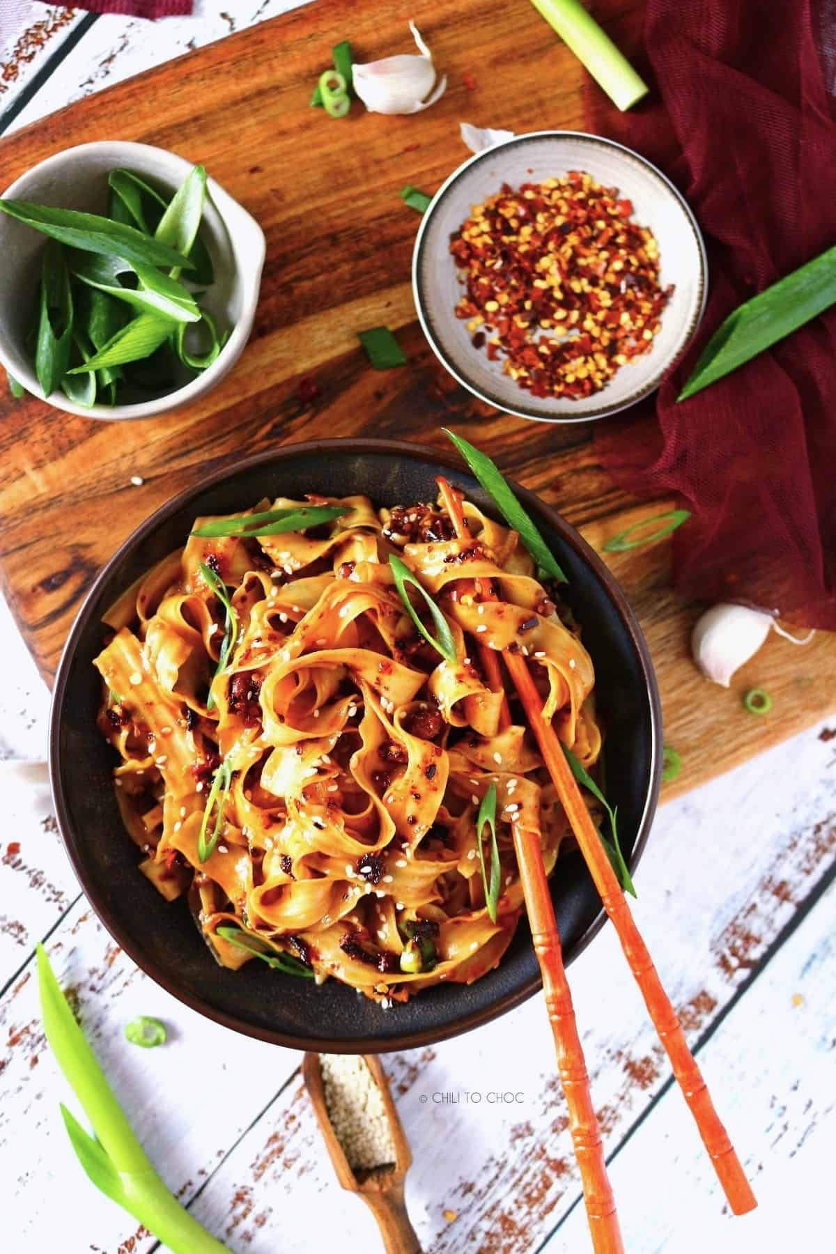 A dish of chili noodles with a bowl of green onions and another small plate of chili flakes.