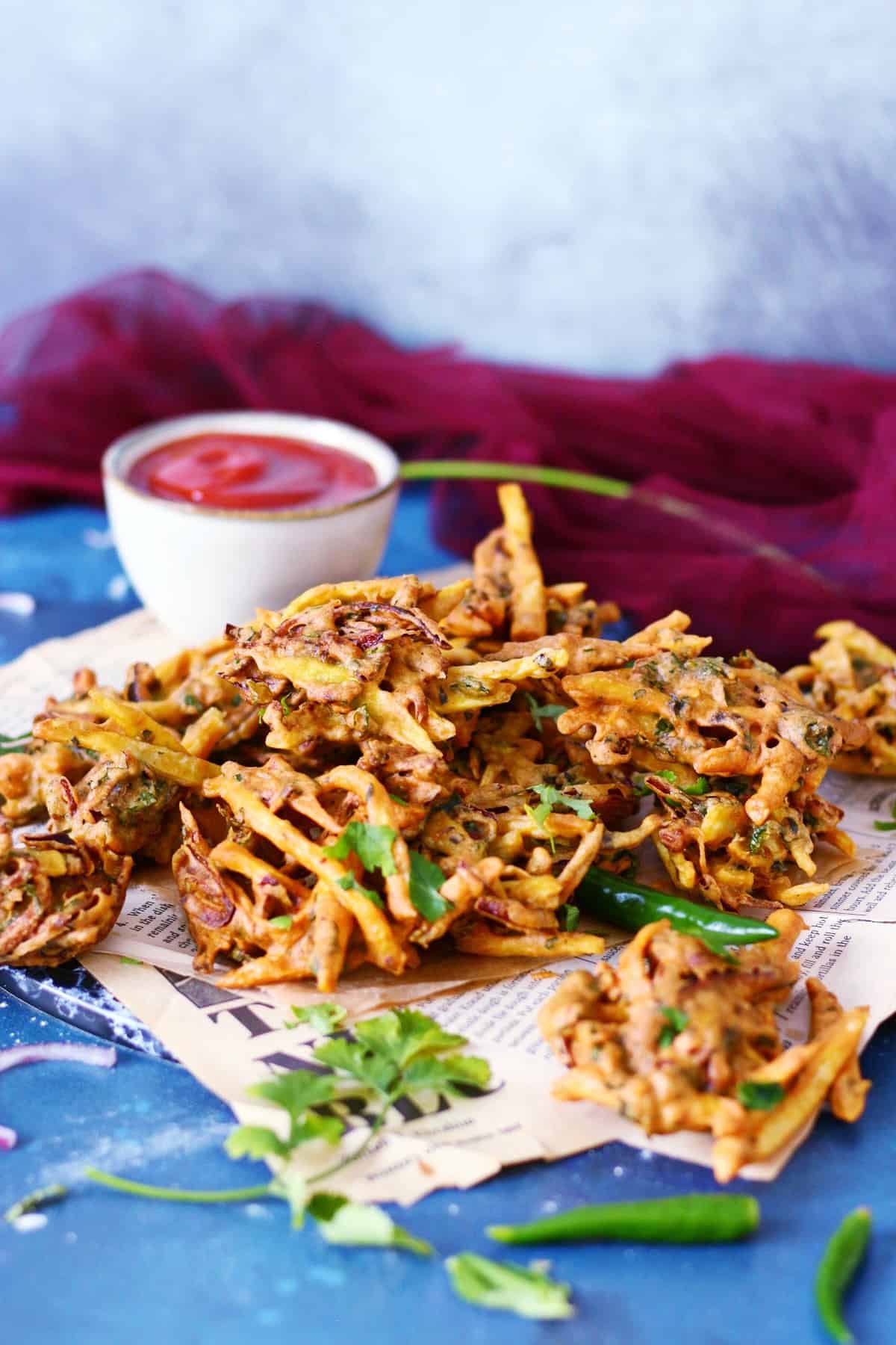Golden brown pakoras on a newspaper with a burgundy fabric behind them.