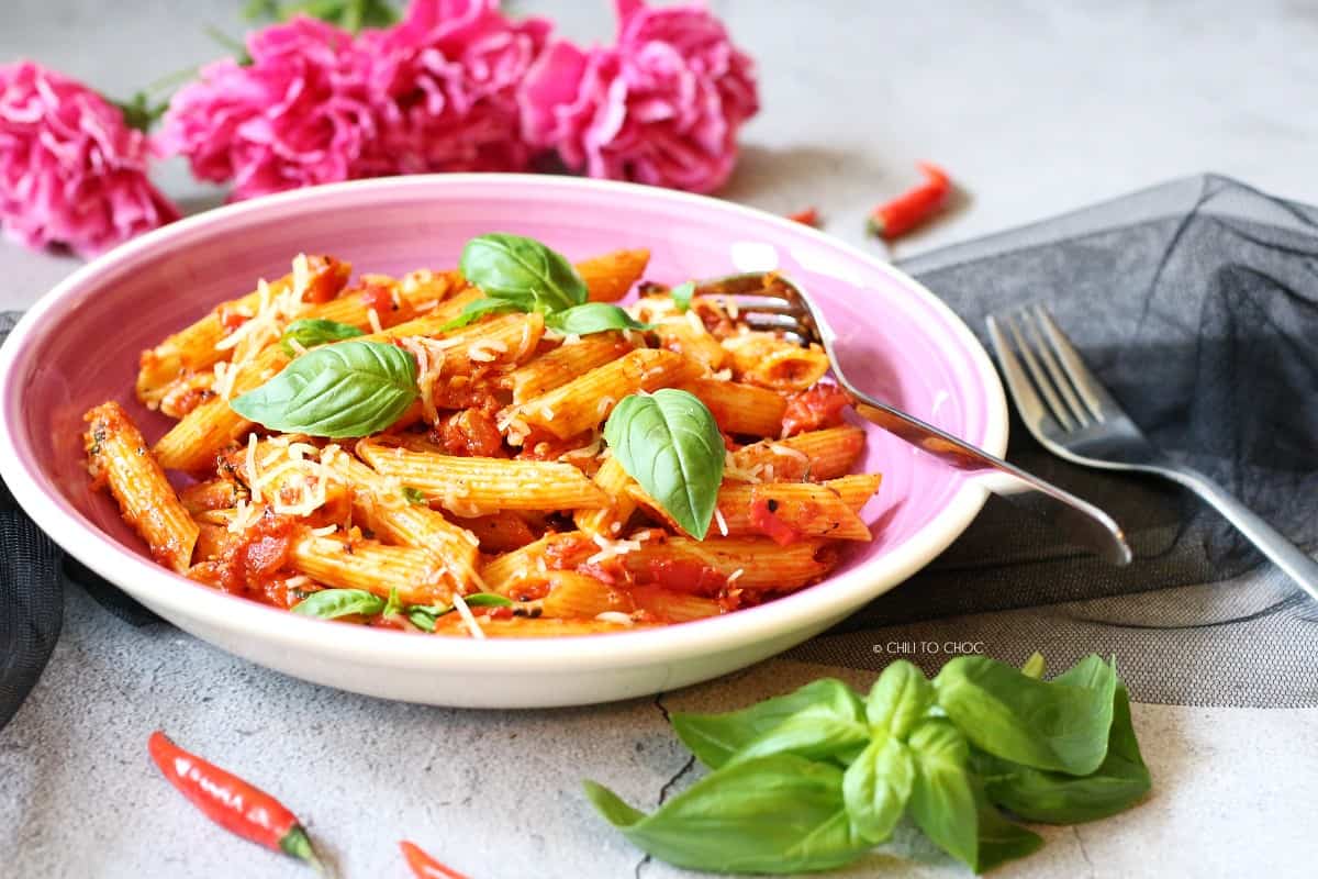 A plate of Pasta Arrabbiata surrounded by forks, flowers, fresh basil and red chili pepper.