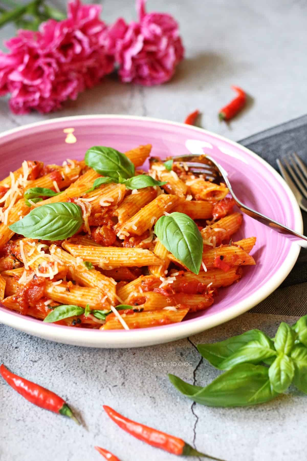Penne Arrabbiata in a pink dish with fresh basil leaves and whole red chili peppers around it.