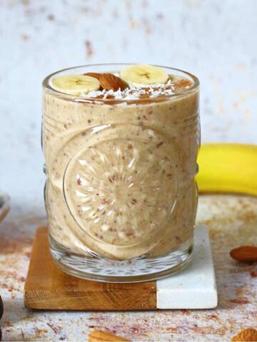 Banana Date Smoothie in a glass, on a coaster with a banana and dates around it.