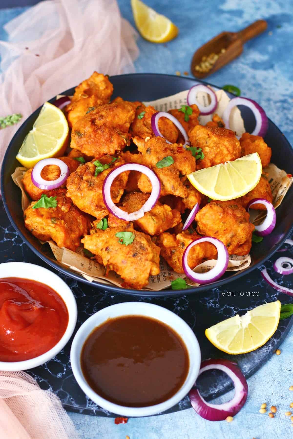 A plate of fish pakoras with side bowls of tamarind chutney and ketchup.