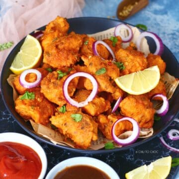 Fish Pakoras on a black dish garnished with onion rings and lemon wedges.