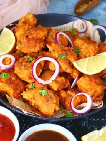 Fish Pakoras on a black dish garnished with onion rings and lemon wedges.
