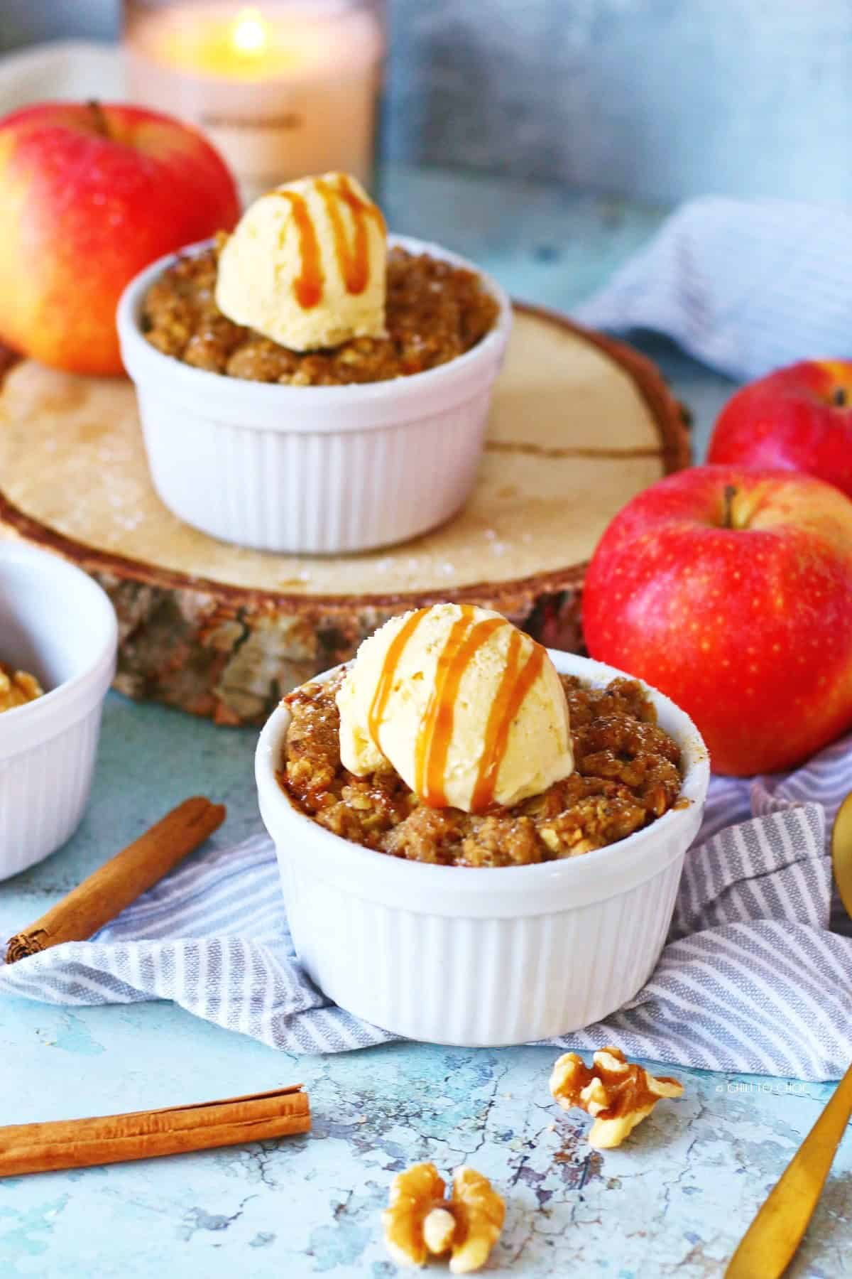 Individual apple crumble in ramekins surrounded by walnuts, cinnamon sticks and whole apples.