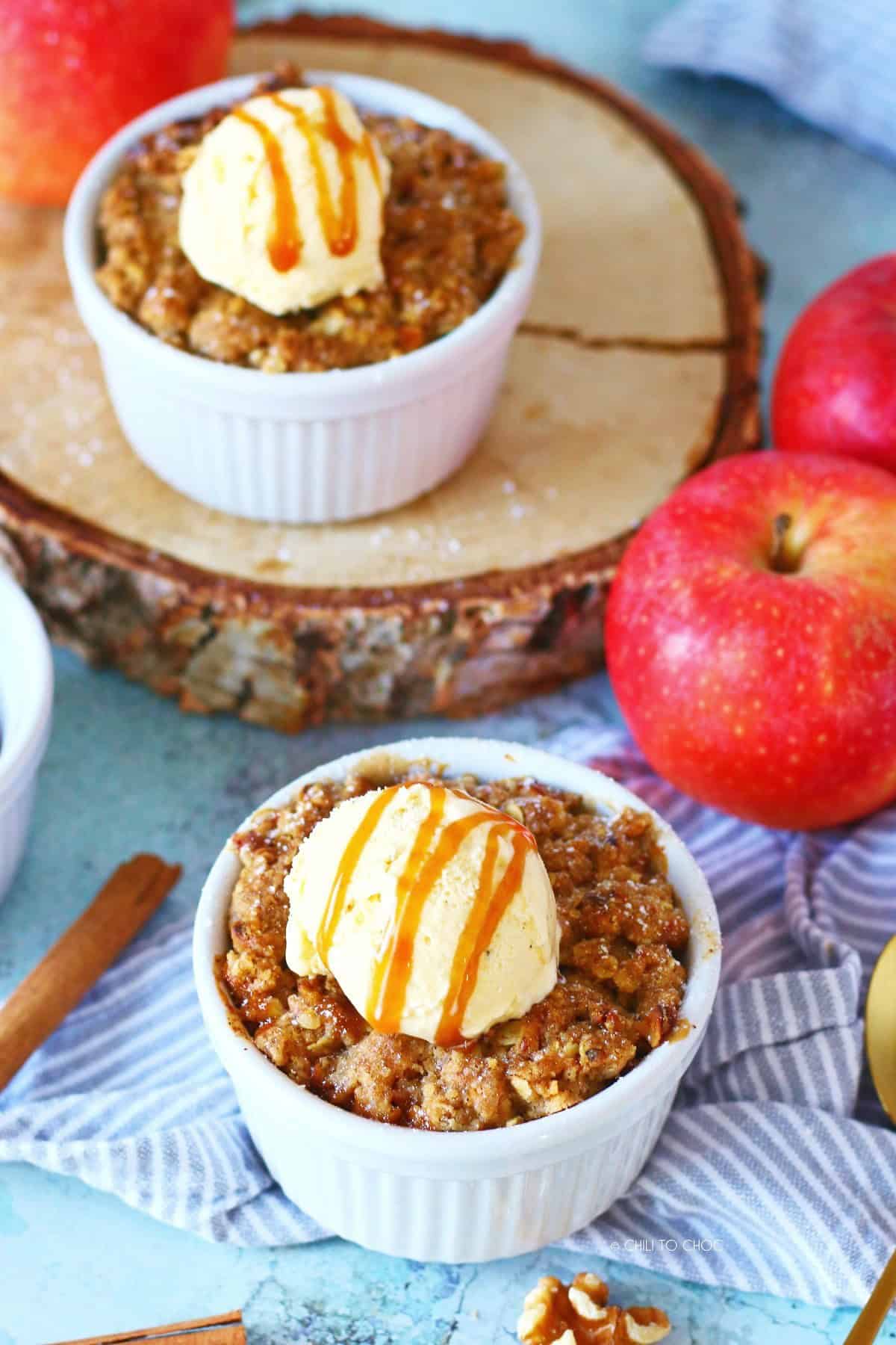 Two mini apple crumble in ramekins with ice cream and top and whole apples on the side.