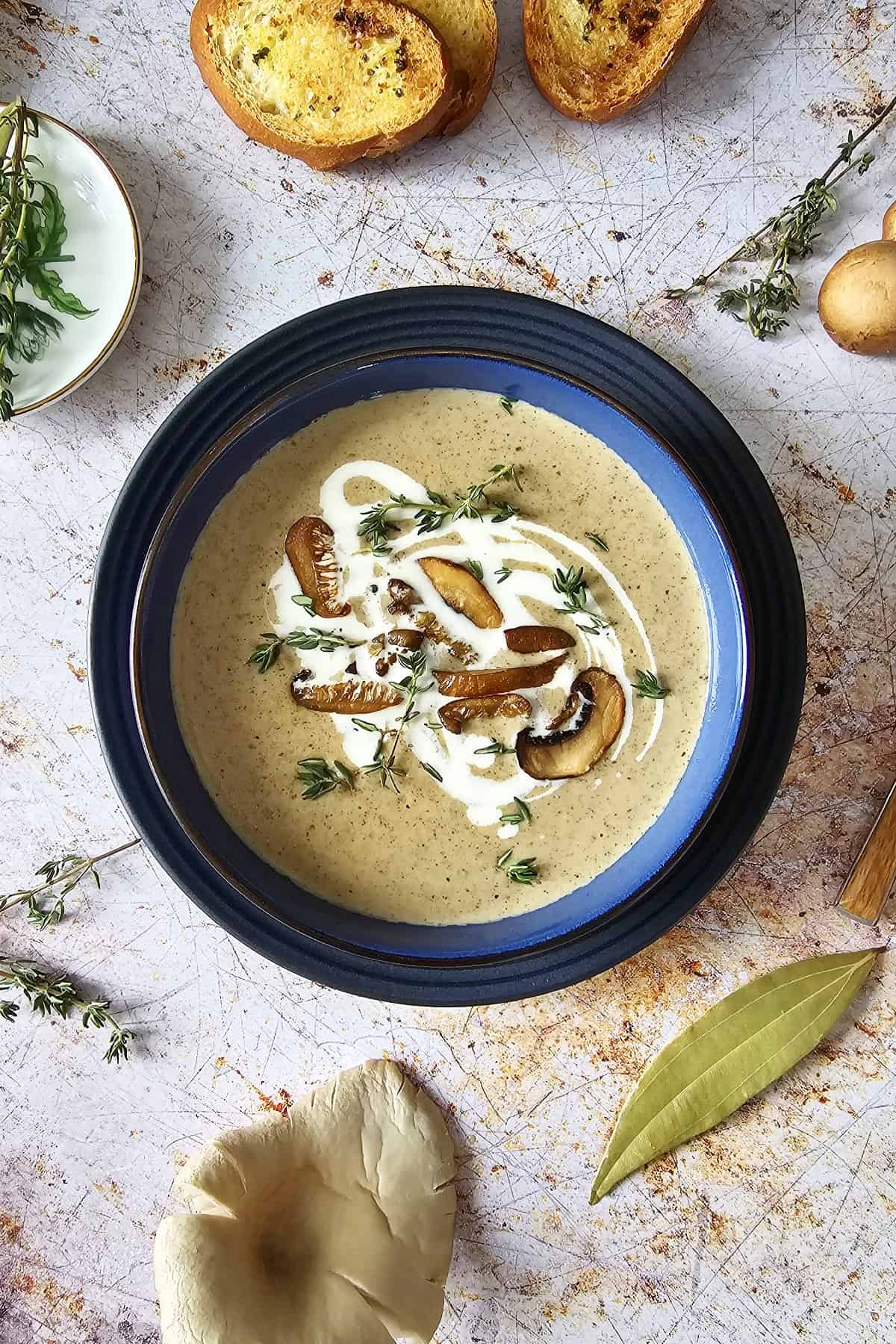 Mushroom soup with garnishes in a blue bowl surrounded by bay leaf, whole mushrooms and thyme sprigs.