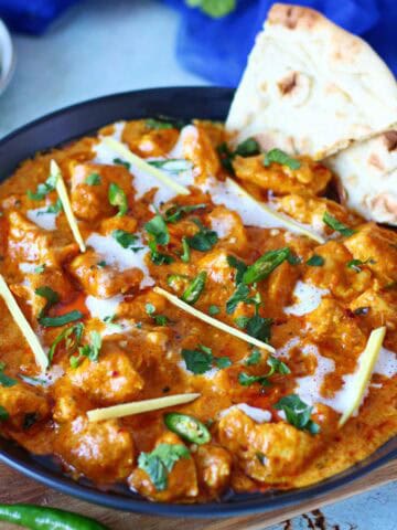 Chicken handi in a black plate garnished with sliced ginger, cilantro, chopped green chilies and naan bits on the side.