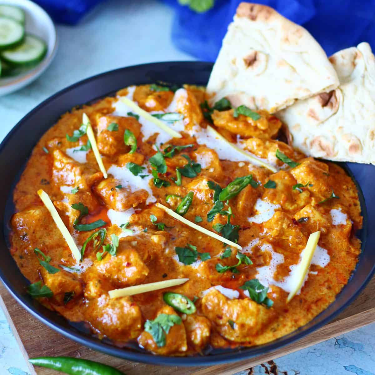 Chicken handi in a black plate garnished with sliced ginger, cilantro, chopped green chilies and naan bits on the side.