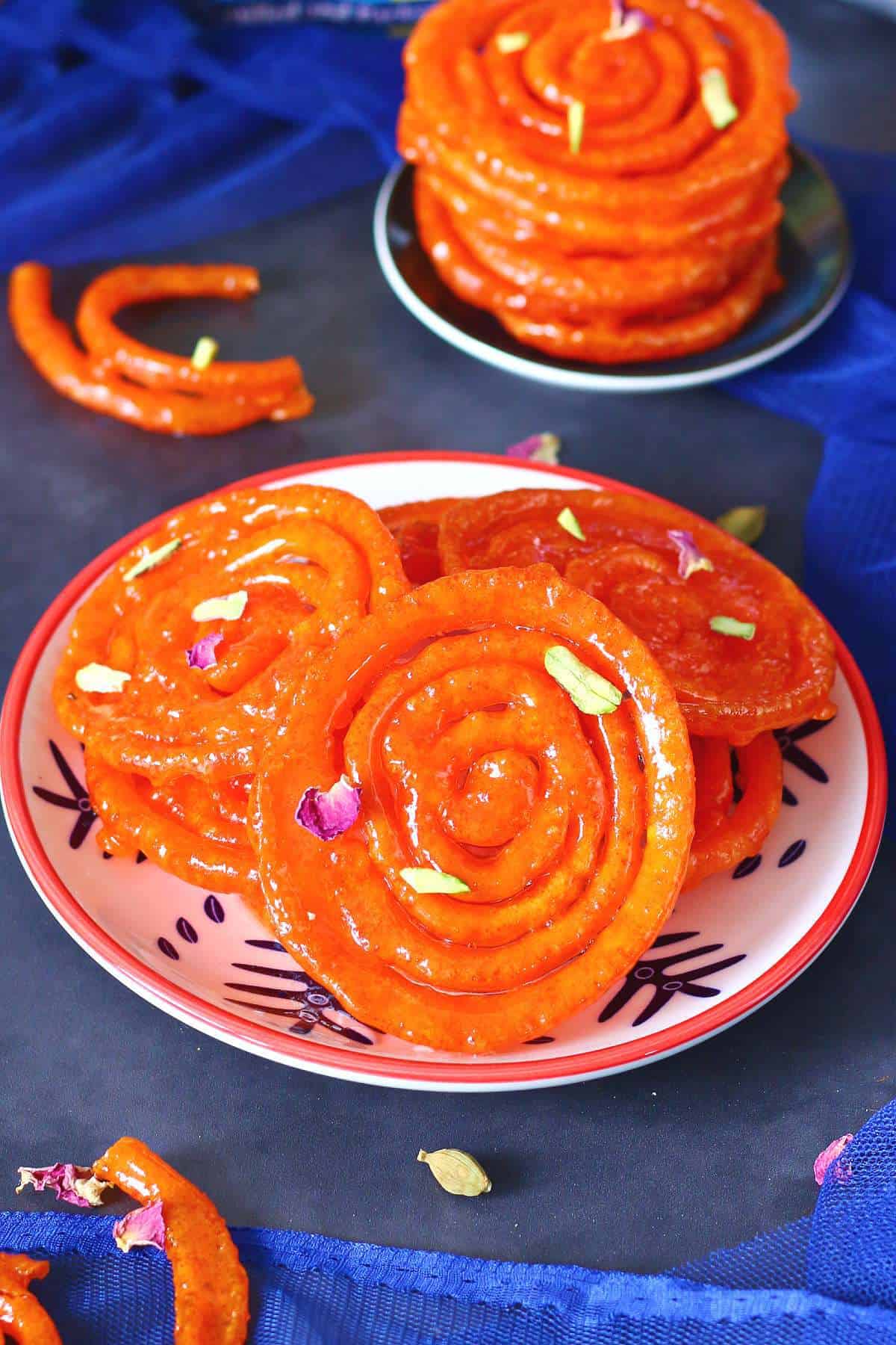 Jalebis on a small plate garnished with dried rose petals and chopped pistachios on a dark background with a stack of jalebis behind.
