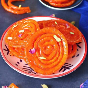 Jalebis on a small plate garnished with dried rose petals and chopped pistachios on a dark background.