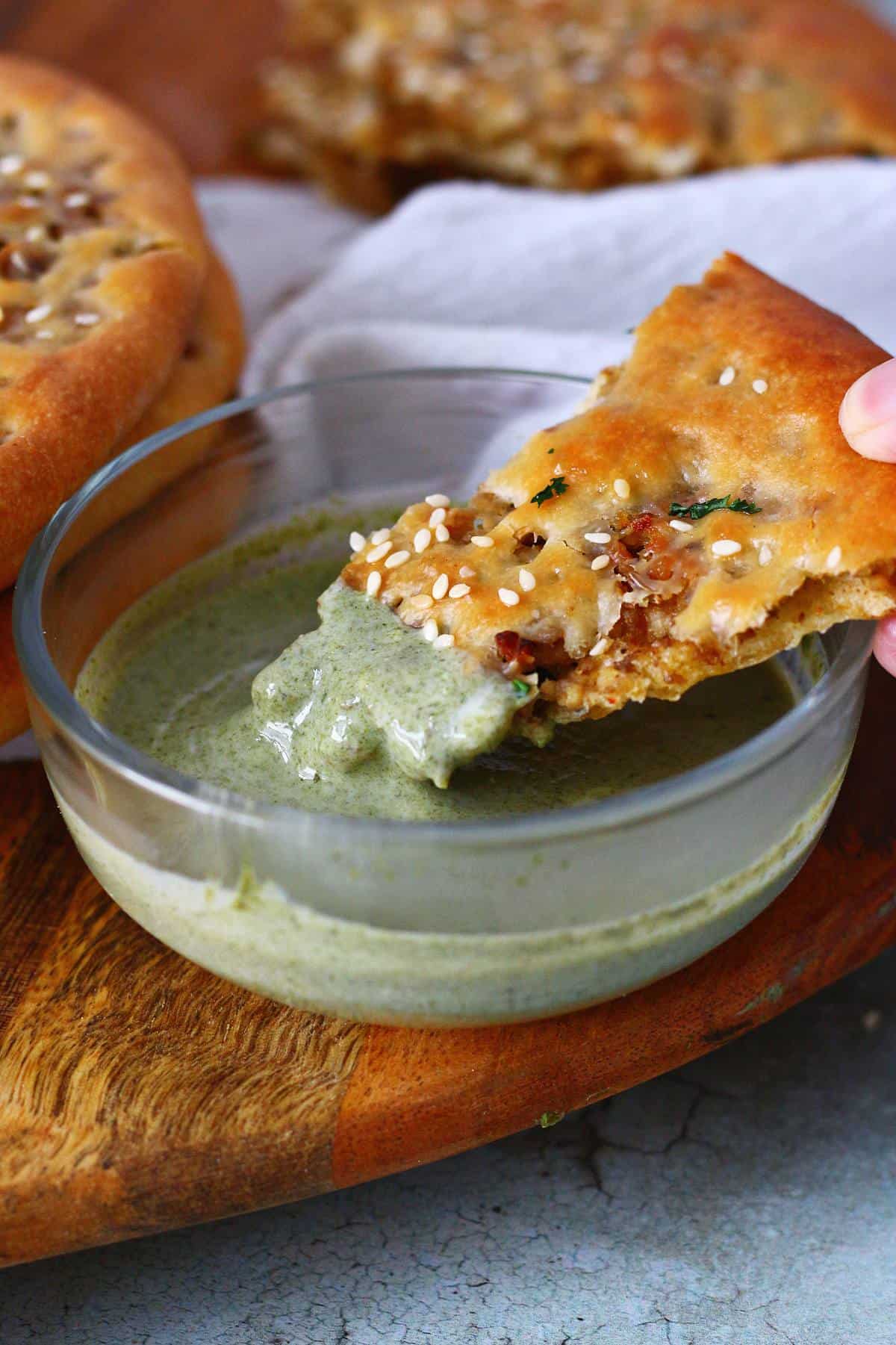 Dipping a piece of keema naan in green raita on a wooden board.