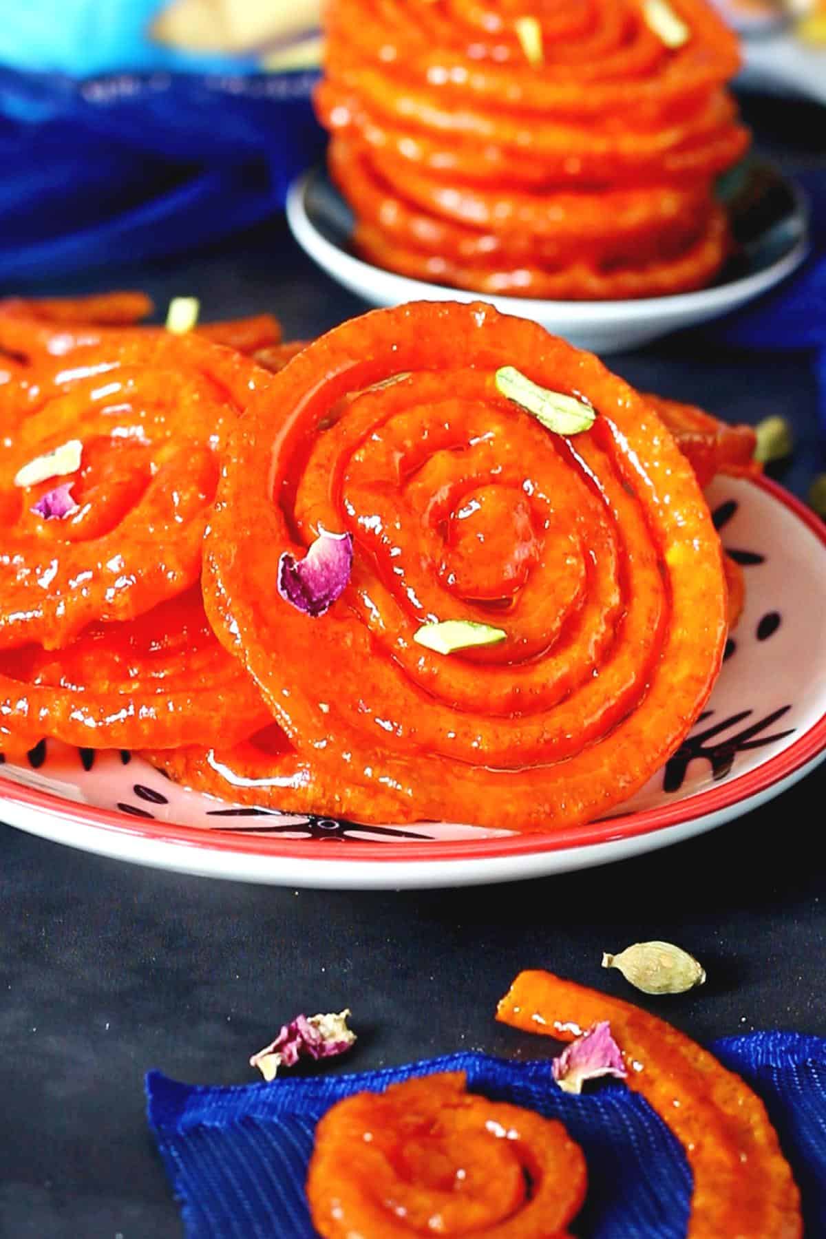 Multiple stacks of jalebi on different plates garnished with rose petals and sliced pistachios.