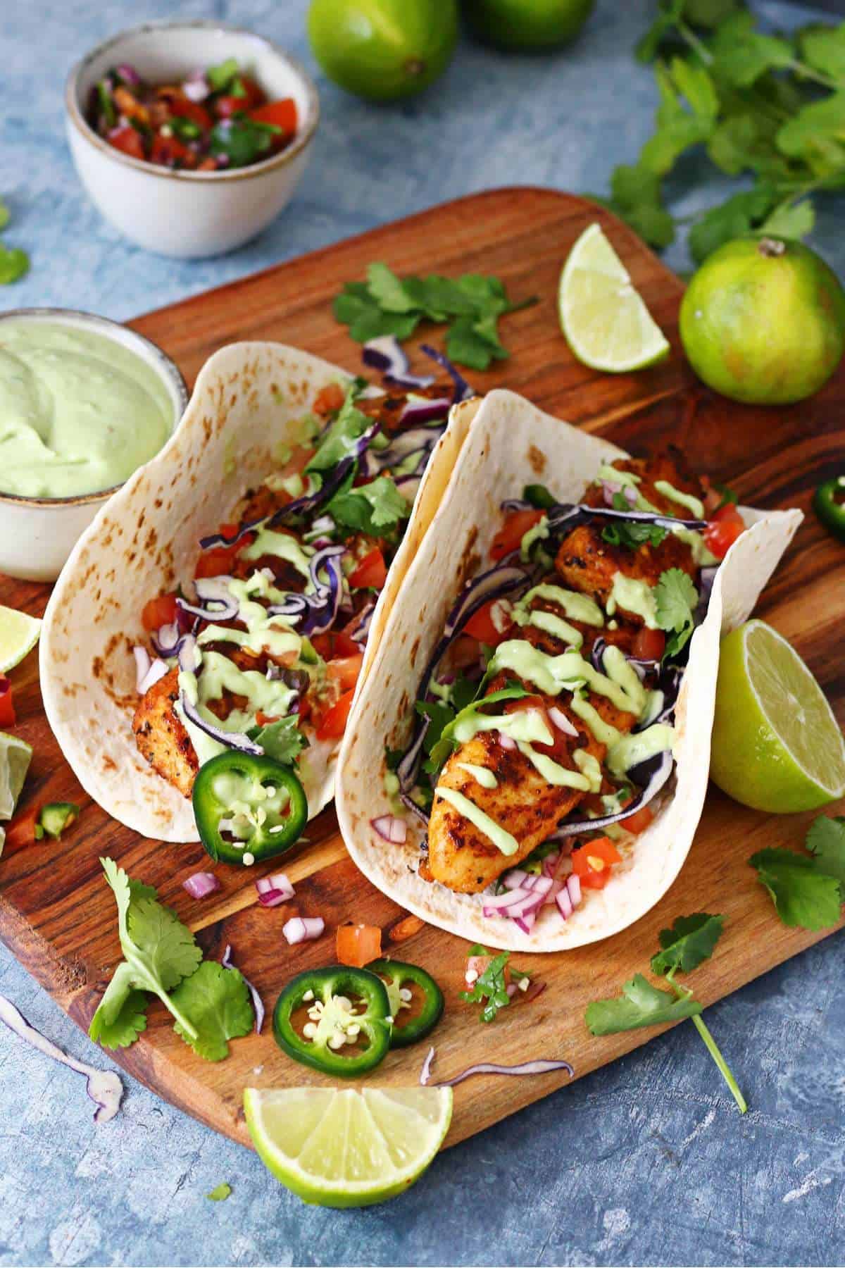 Wooden board with two fish tacos on an aesthetic background with avocado crema and pico de gallo in dip bowls.