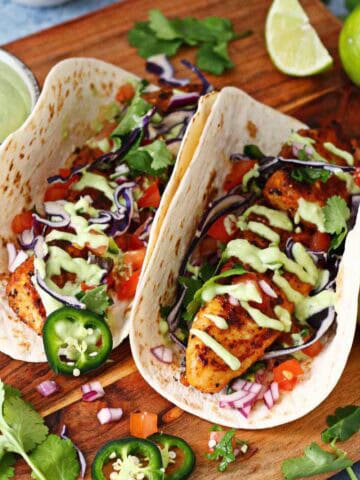 Two blackened fish tacos on a wooden board with garnishes around it.