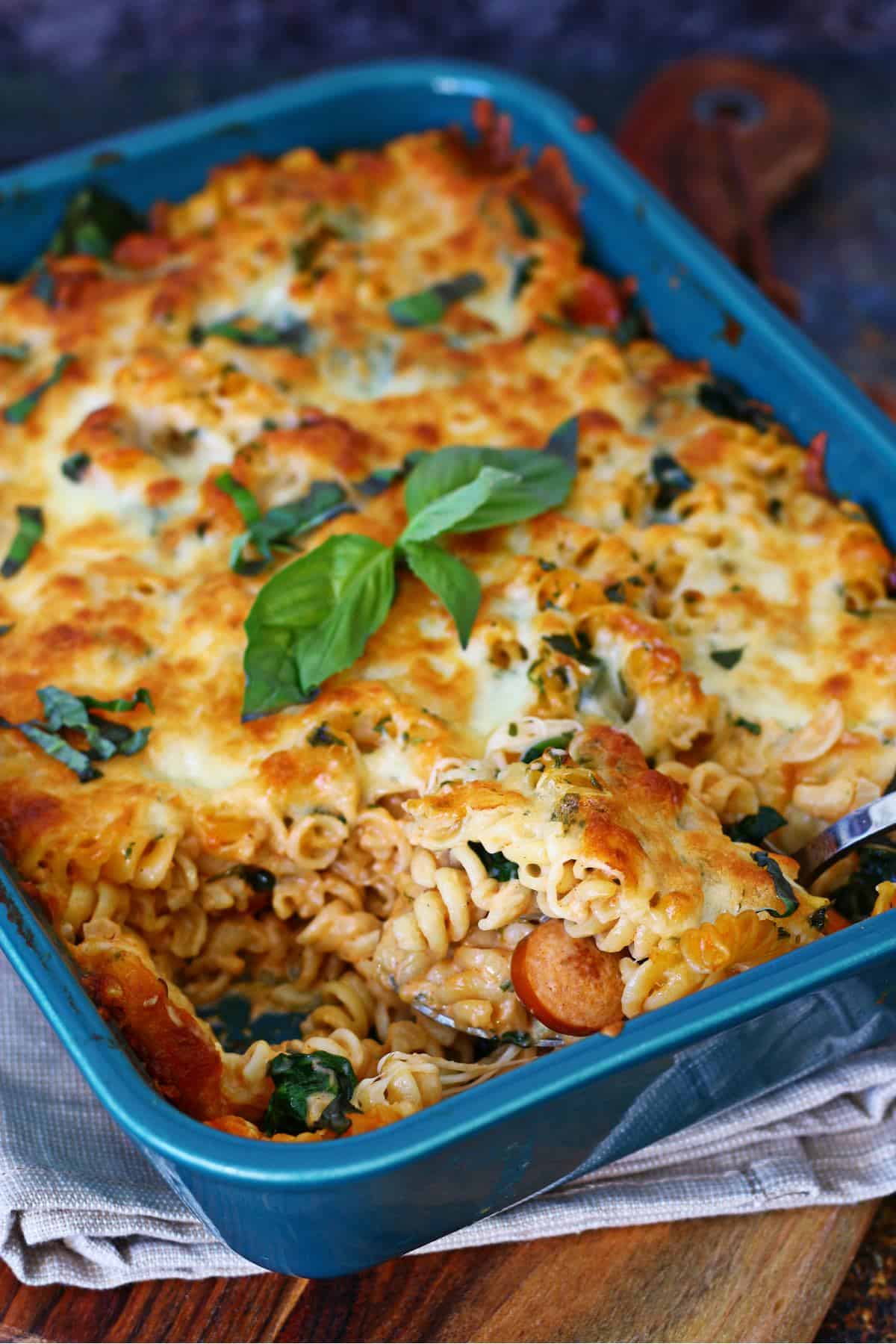 Scooping out baked pasta with sausages from a casserole dish.