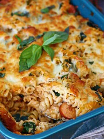 Scooping out baked sausage pasta from a blue casserole dish with fresh basil garnish.