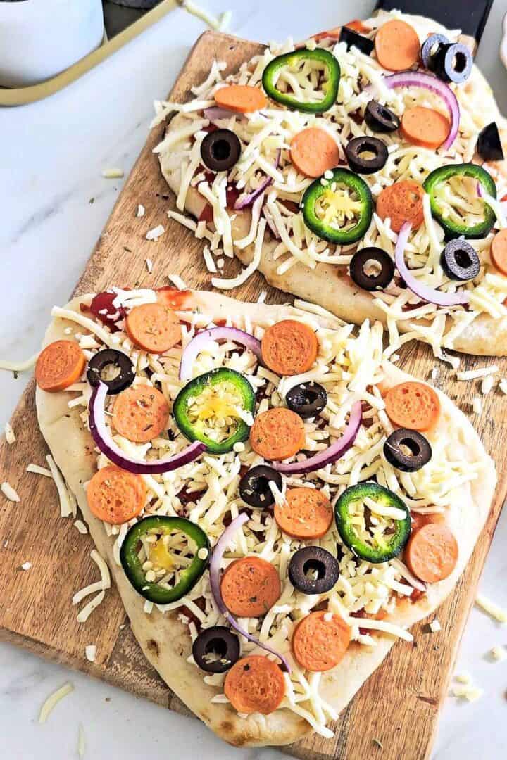 Two flatbreads on a wooden board topped with pizza sauce, cheese, pepperoni, black olives and sliced jalapenos.