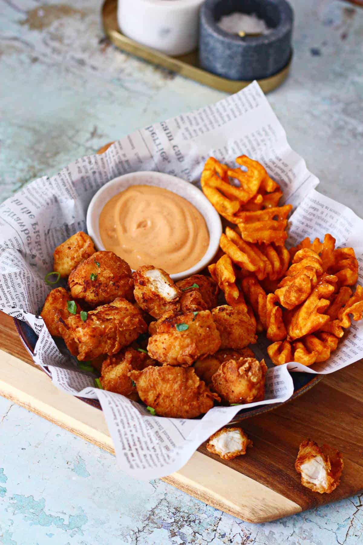 Chicken nuggets in a shallow bowl with waffles fries and chipotle dip on a wooden board with salt and pepper at the back.