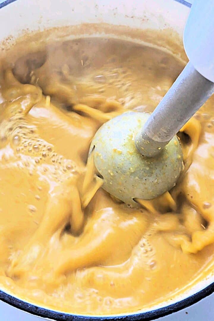 An immersion blender blending the lentil soup in a white pot.