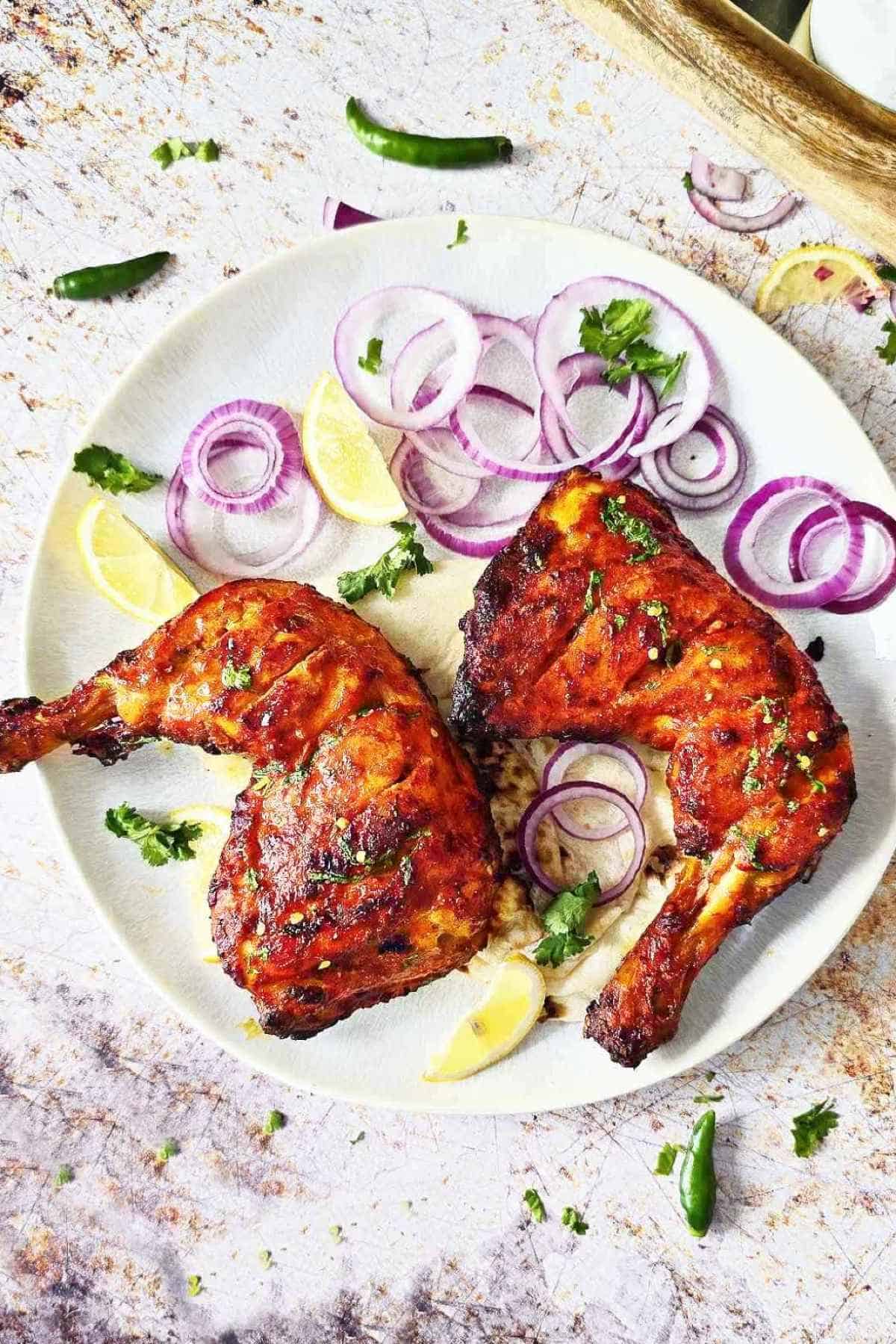 Top shot of a plate of tandoori chicken with sides, surrounded by whole green chilies and chopped cilantro.
