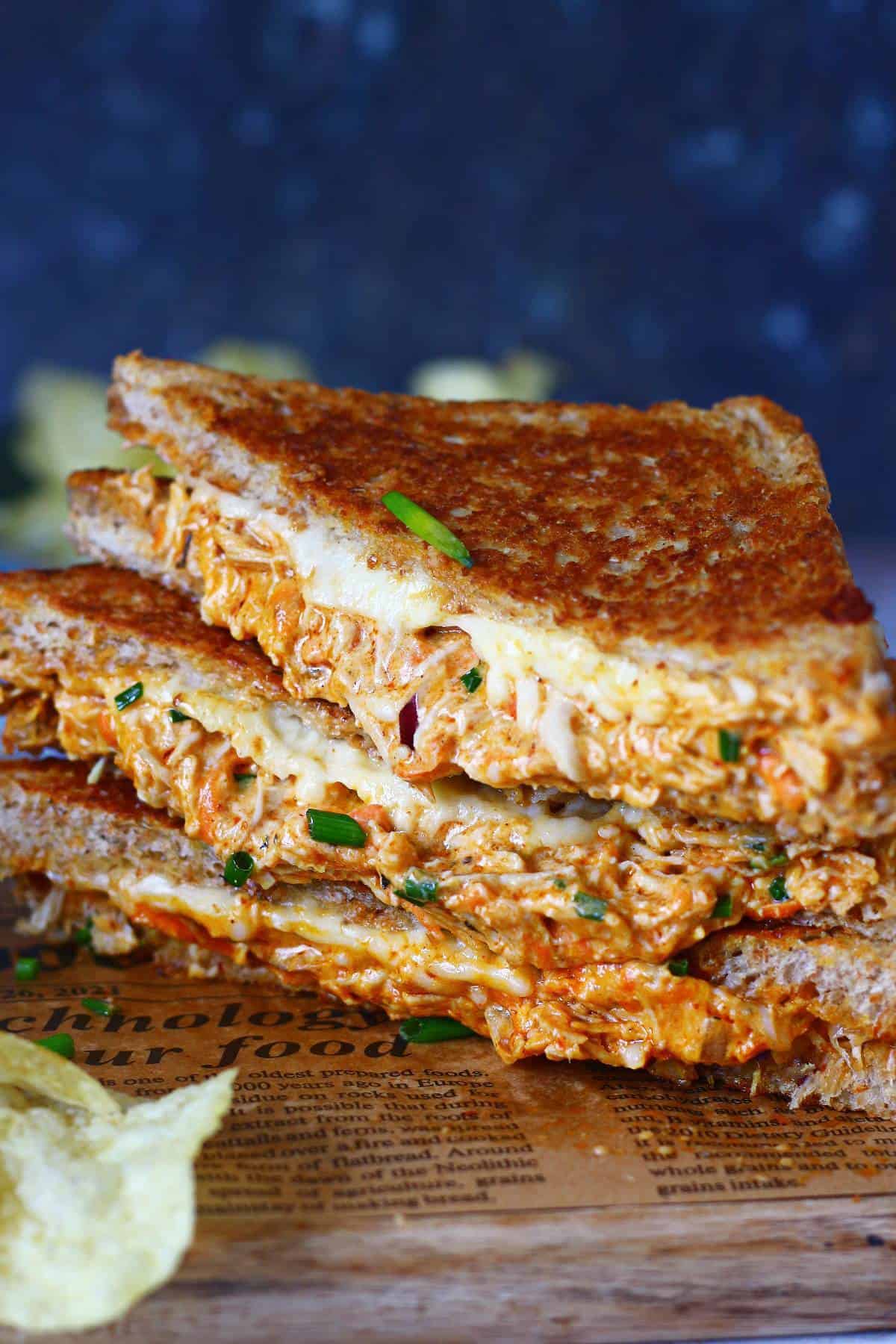 A stack of chicken melt triangles on a brown paper with a piece of crisp in the front.
