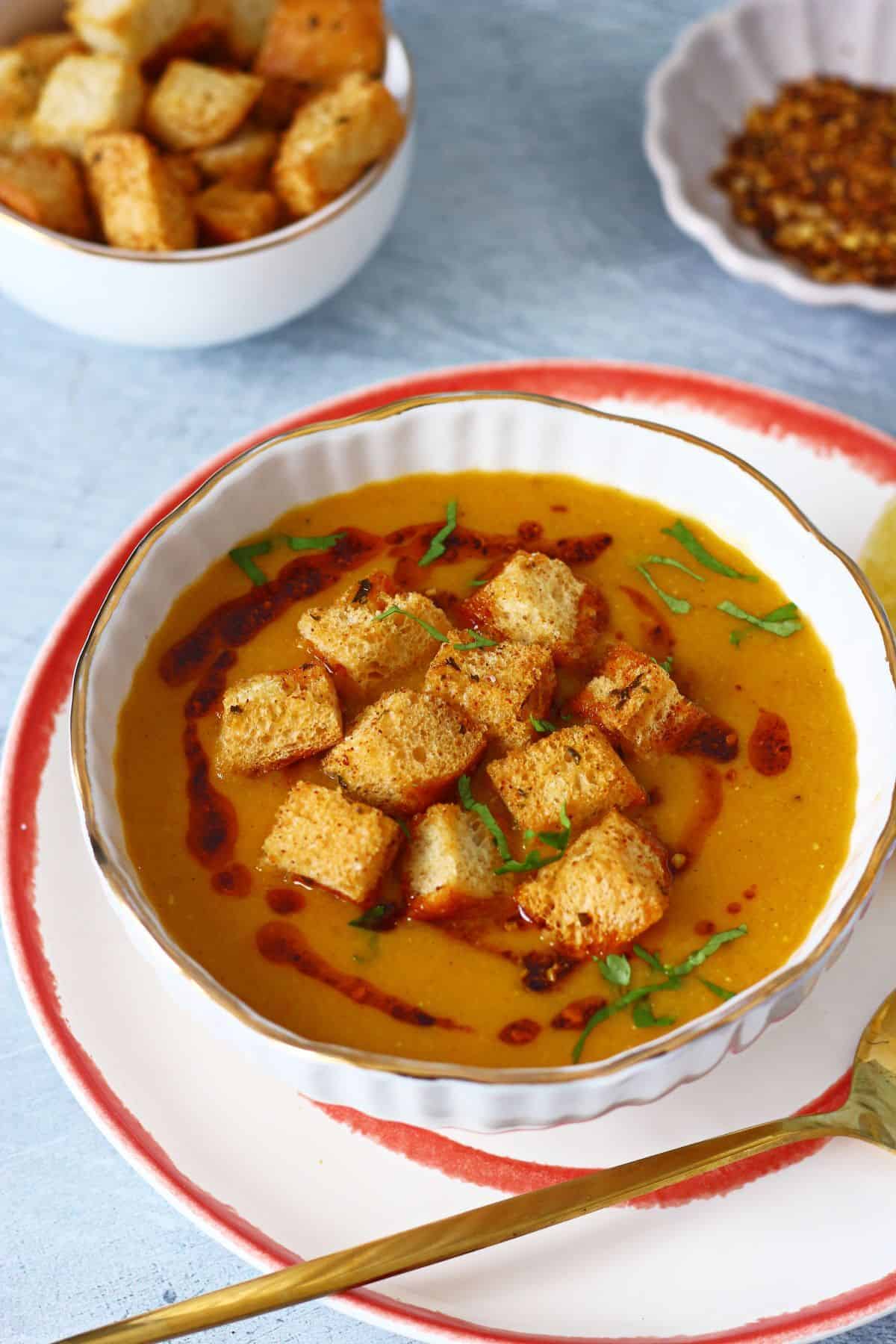A bowl of Turkish red lentil soup served on an orange-rimmed plate with a gold spoon, croutons, and red pepper flakes in the background.