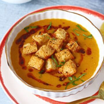 Red lentil soup in a white bowl topped with croutons, chili oil and fresh chopped cilantro.