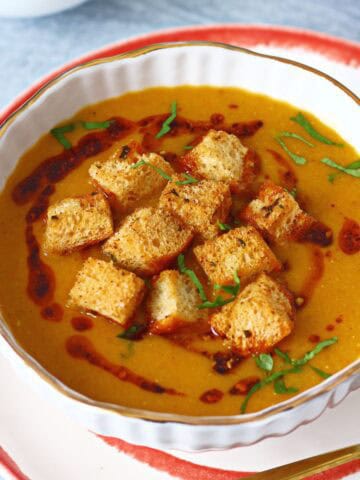 Red lentil soup in a white bowl topped with croutons, chili oil and fresh chopped cilantro.