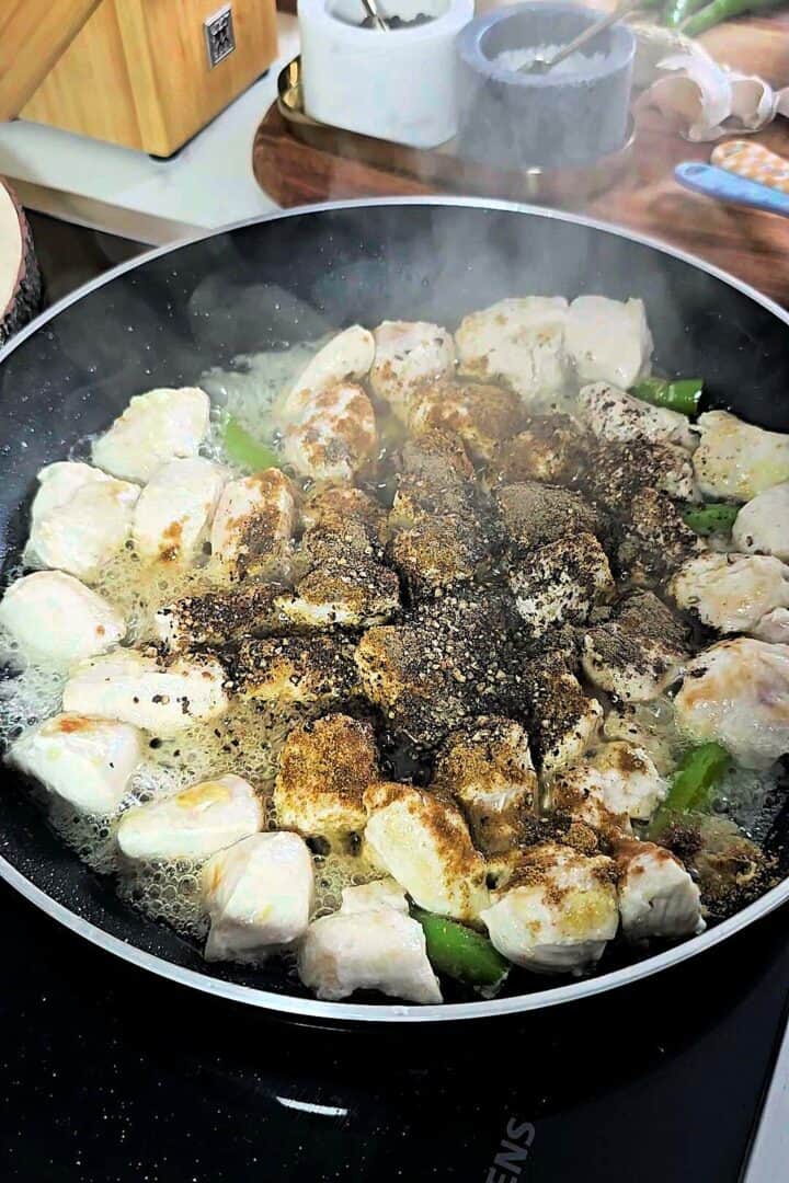 Ground black pepper and spices added to sautéed chicken pieces in the black pan.