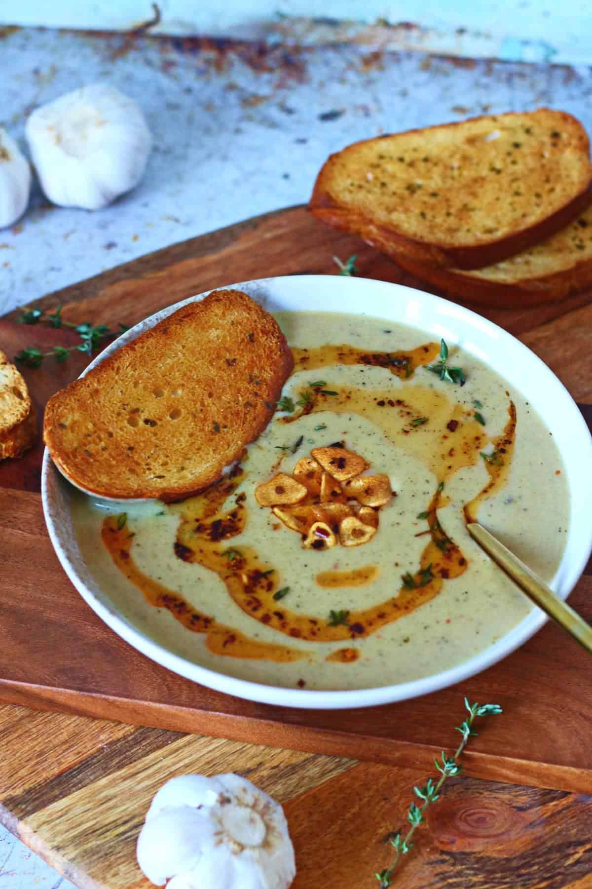 A bowl of roasted garlic soup topped with garlic chips and chili oil, served with toasted bread.