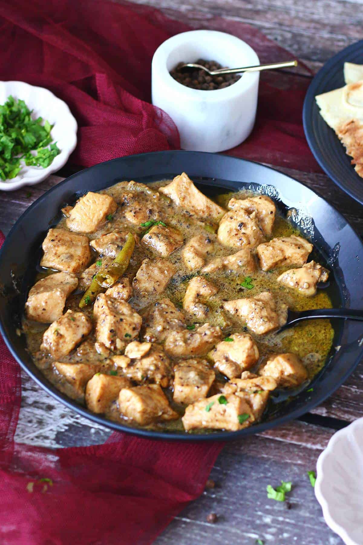 Chicken Kali Mirch served in a black bowl with roti, fresh cilantro, and black peppercorns surrounding it.