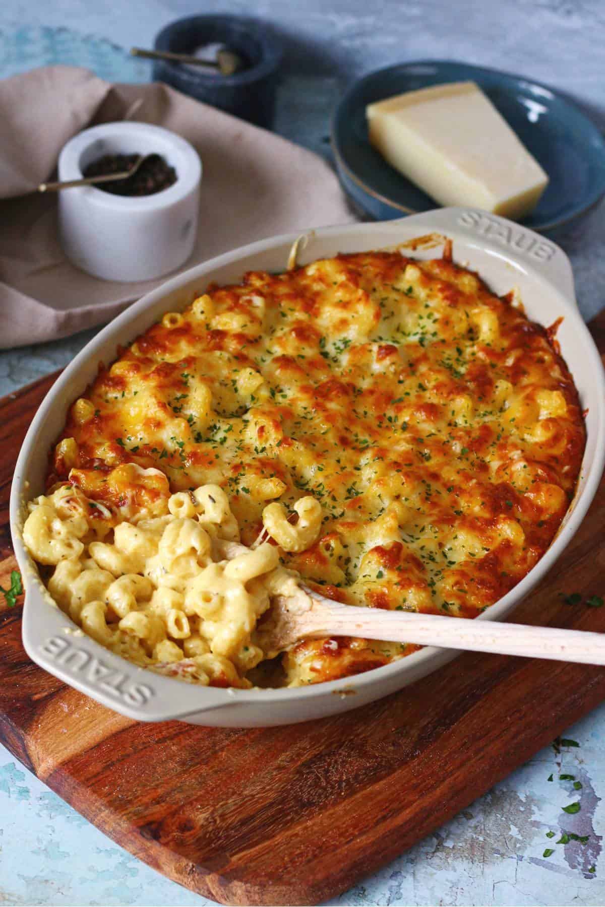 Full shot of the Baked Four Cheese Mac & Cheese recipe in a stoneware casserole dish, with Parmesan cheese block visible in the background.